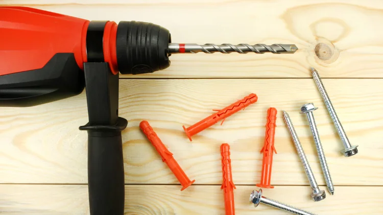 Hammer drill with masonry bit, plastic wall anchors, and metal screws on wooden surface